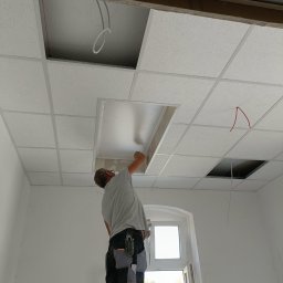 Ogolno budowlana - Montaż sufitu podwieszanego kasetonowego w pomieszczeniu z oknem; widoczne otwory na oświetlenie i instalacje, pracownik na drabinie w ubraniu roboczym.