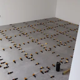 Szare płytki podłogowe w trakcie układania, z użyciem systemu poziomowania płytek z czarnymi klinami i żółtymi podstawkami, widoczna poziomica SLOWIK.