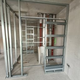 Ogolno budowlana - Metalowa konstrukcja szkieletowa ścian działowych w surowym stanie, widoczne instalacje wentylacyjne i elektryczne w tle, przygotowane do dalszych prac wykończeniowych.