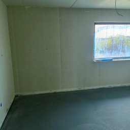 Ogolno budowlana - Surowe wnętrze w trakcie wykańczania: ściany i sufit pokryte płytami, zabezpieczone okno z widokiem na zieleń, wylewka na podłodze.