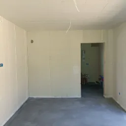 Surowe wnętrze w trakcie remontu: ściany wyłożone płytami gipsowo-kartonowymi, surowa wylewka na podłodze, widoczne instalacje w sąsiednim pomieszczeniu przez otwór drzwiowy.