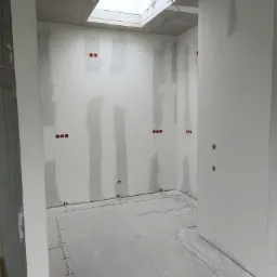 Wnętrze w trakcie prac wykończeniowych: ściany z widocznymi łączeniami płyt, zamontowane puszki elektryczne oraz kable ułożone na podłodze, widoczne okno dachowe.