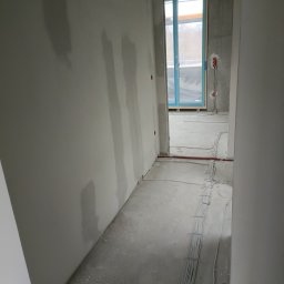 Ogolno budowlana - Korytarz w surowym stanie, widoczne przewody elektryczne ułożone wzdłuż ściany, ściany przygotowane pod malowanie, widoczne ślady po szpachlowaniu.