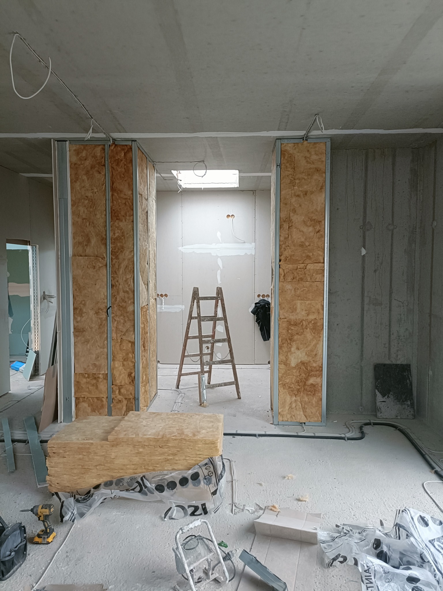 Montaż ścian działowych: widok na szkielet z profili metalowych wypełniony wełną mineralną, częściowo obłożony płytami. Na podłodze leżą paczki wełny i narzędzia, w tle drabina oparta o ścianę.