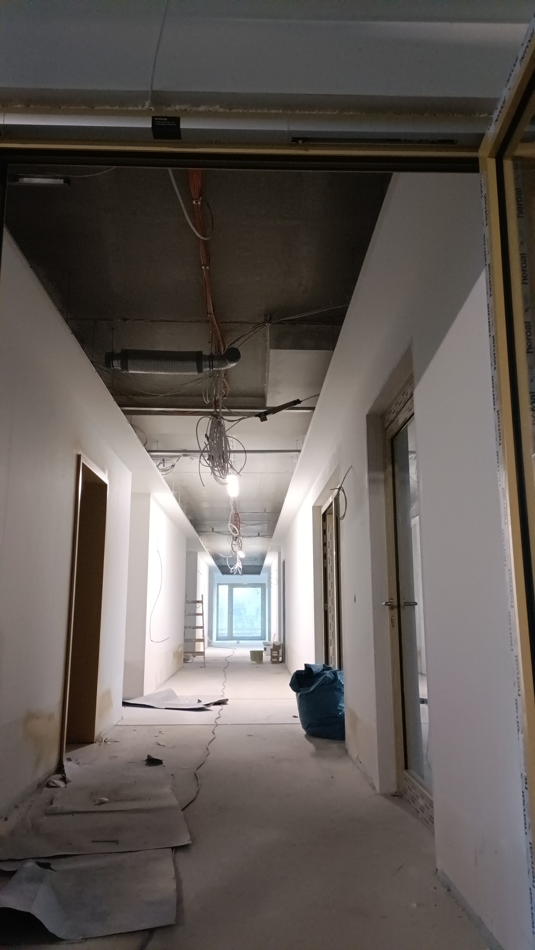 Korytarz biurowy w trakcie prac wykończeniowych: widoczne kable, instalacje pod sufitem, gładkie, białe ściany, resztki materiałów budowlanych na podłodze, drabina w oddali.