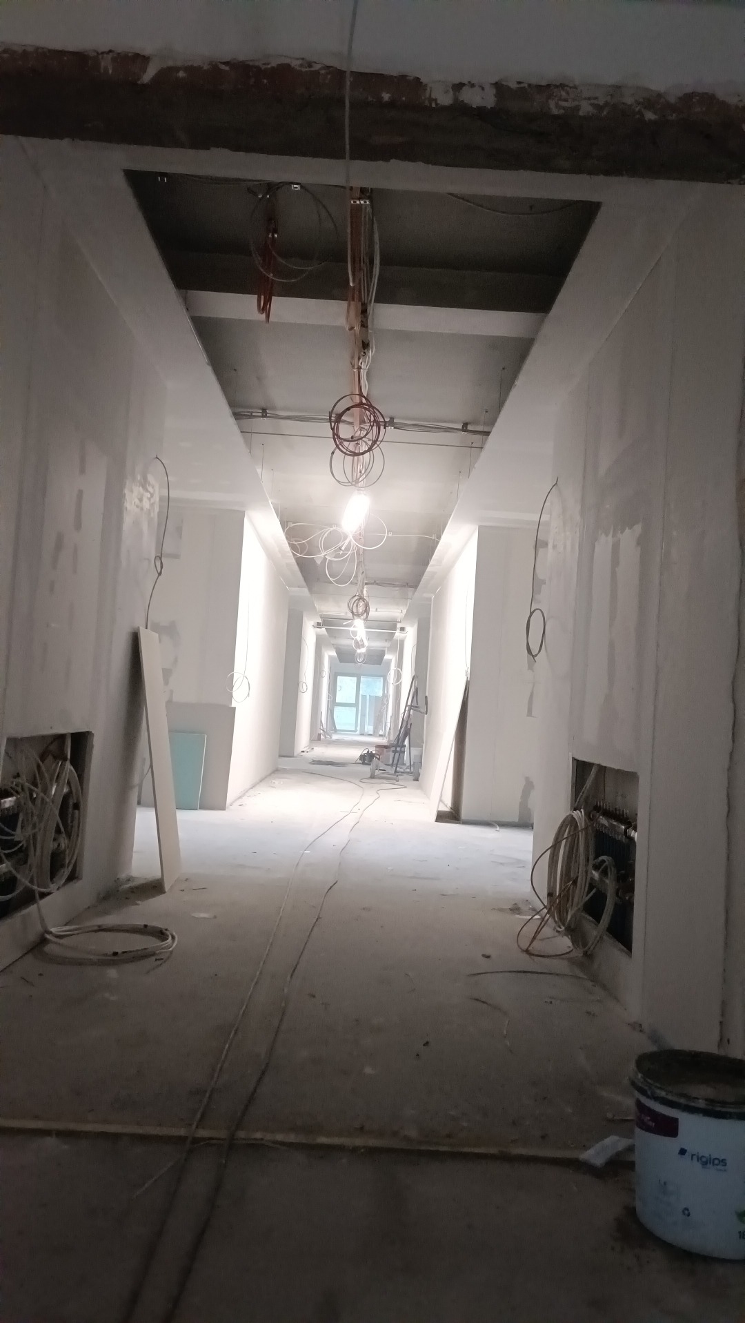 Długi korytarz w trakcie prac wykończeniowych, widoczne otwory w ścianach z instalacjami, ściany przygotowane pod malowanie, surowy betonowy sufit z wiszącymi kablami oświetleniowymi, puszka...