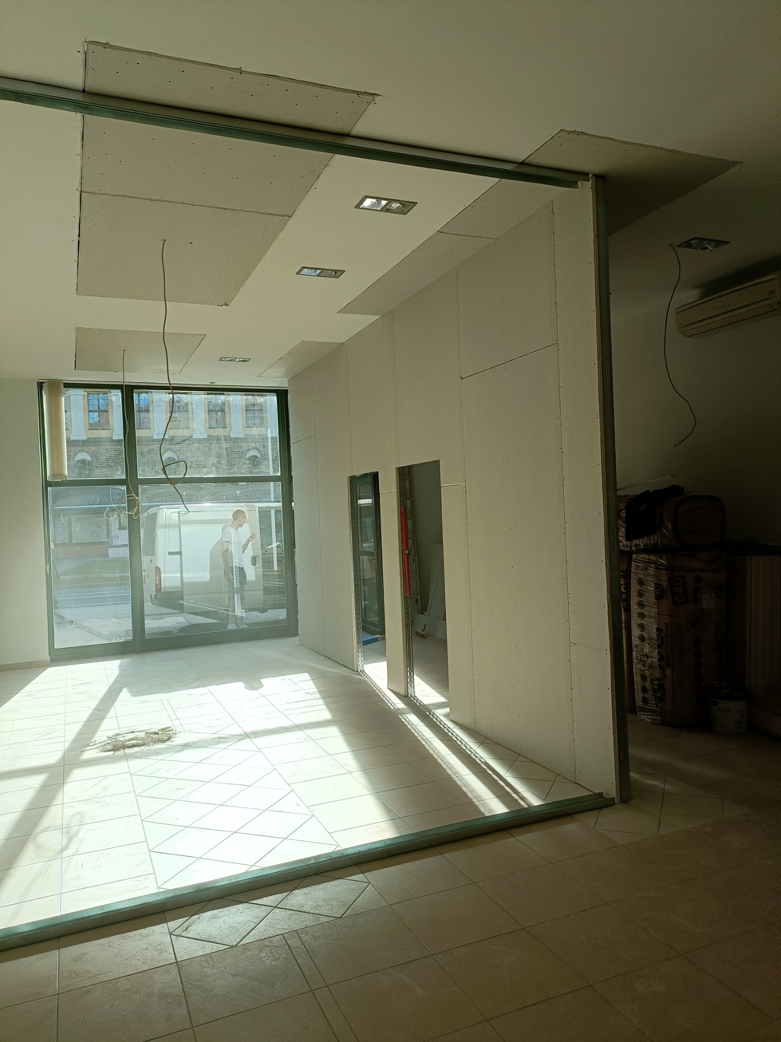 Wnętrze w trakcie montażu ścianki działowej z płyty gipsowo-kartonowej, widoczne profile metalowe i częściowo zamontowane płyty, zarys otworu drzwiowego, w tle okno z widokiem na ulicę i pracownika.