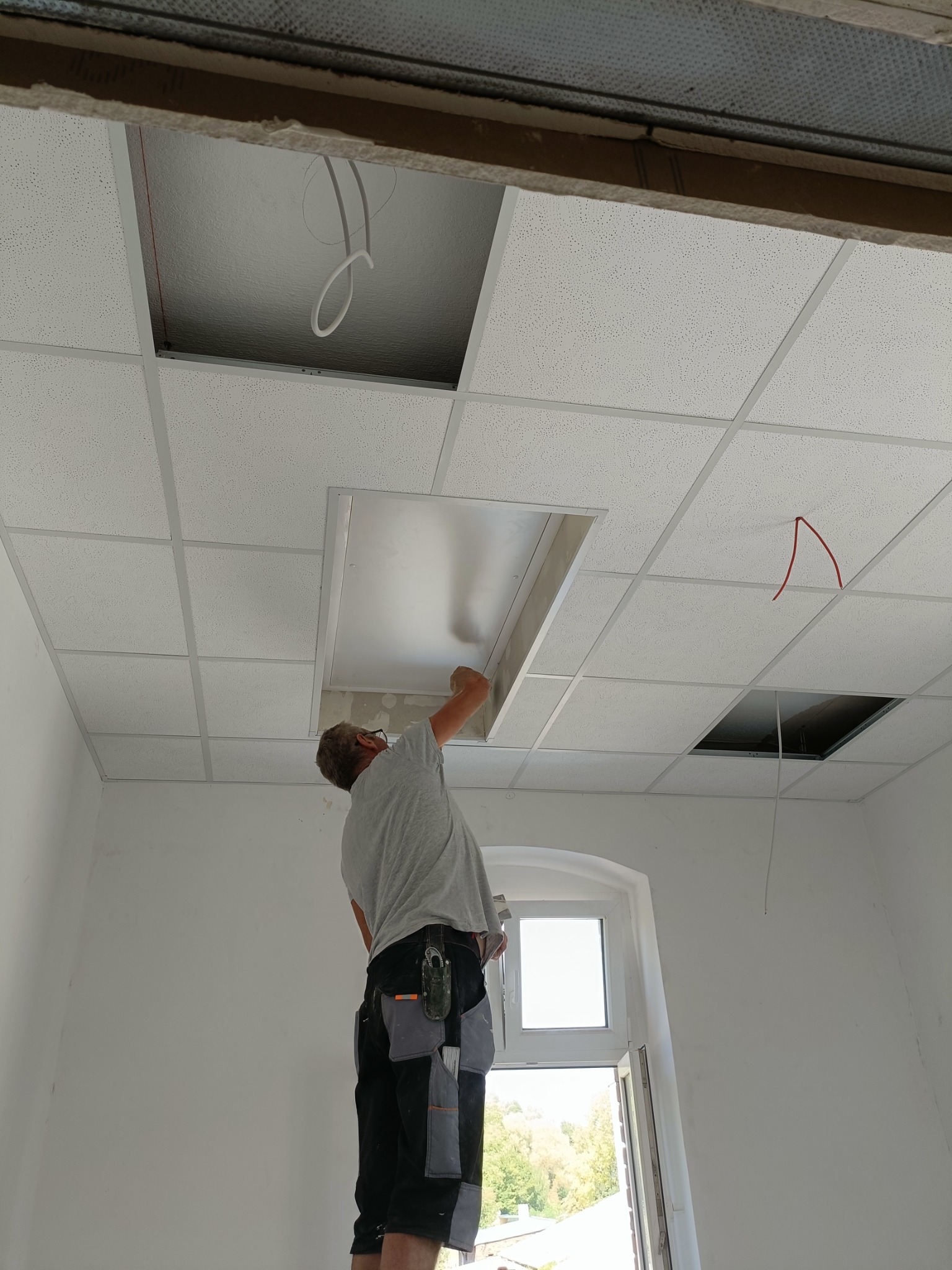 Montaż sufitu podwieszanego kasetonowego w pomieszczeniu z oknem; widoczne otwory na oświetlenie i instalacje, pracownik na drabinie w ubraniu roboczym.