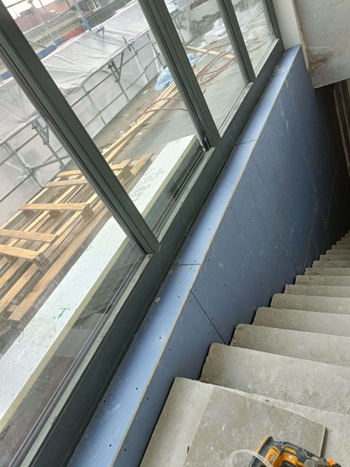 Montaż niebieskich płyt na ścianie przy schodach w nowym budynku, widoczne okno i narzędzia.