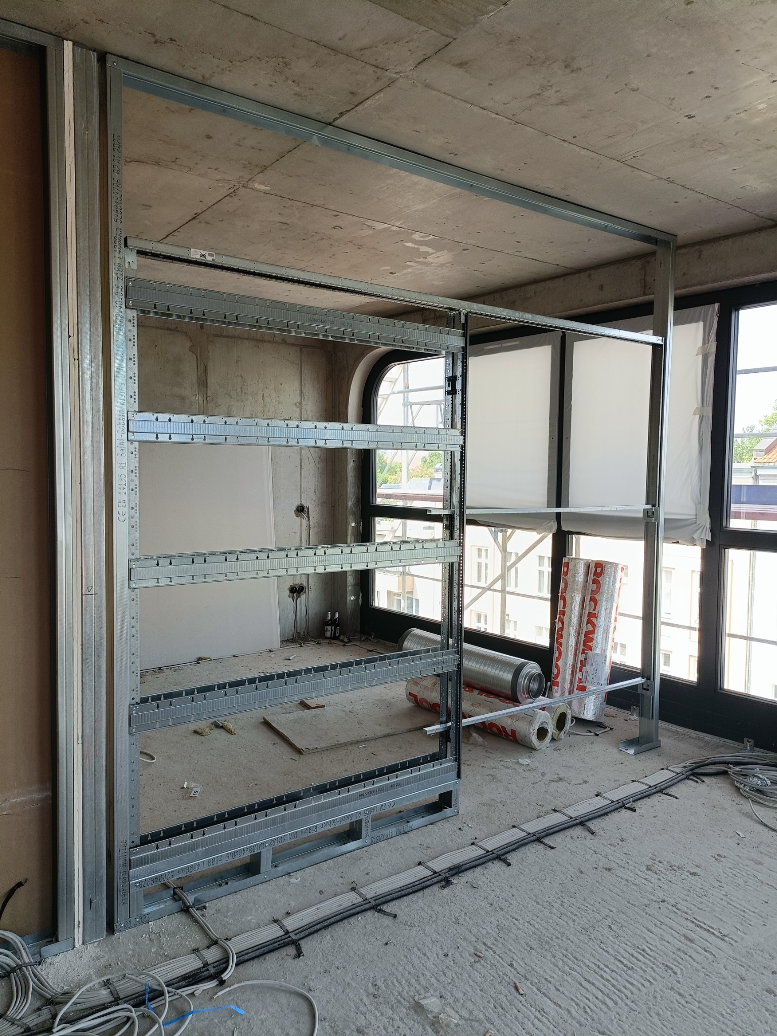 Metalowa konstrukcja szkieletowa pod ścianę działową z płyty karton gips, widoczne przewody elektryczne na podłodze i rolki wełny mineralnej przy oknie.
