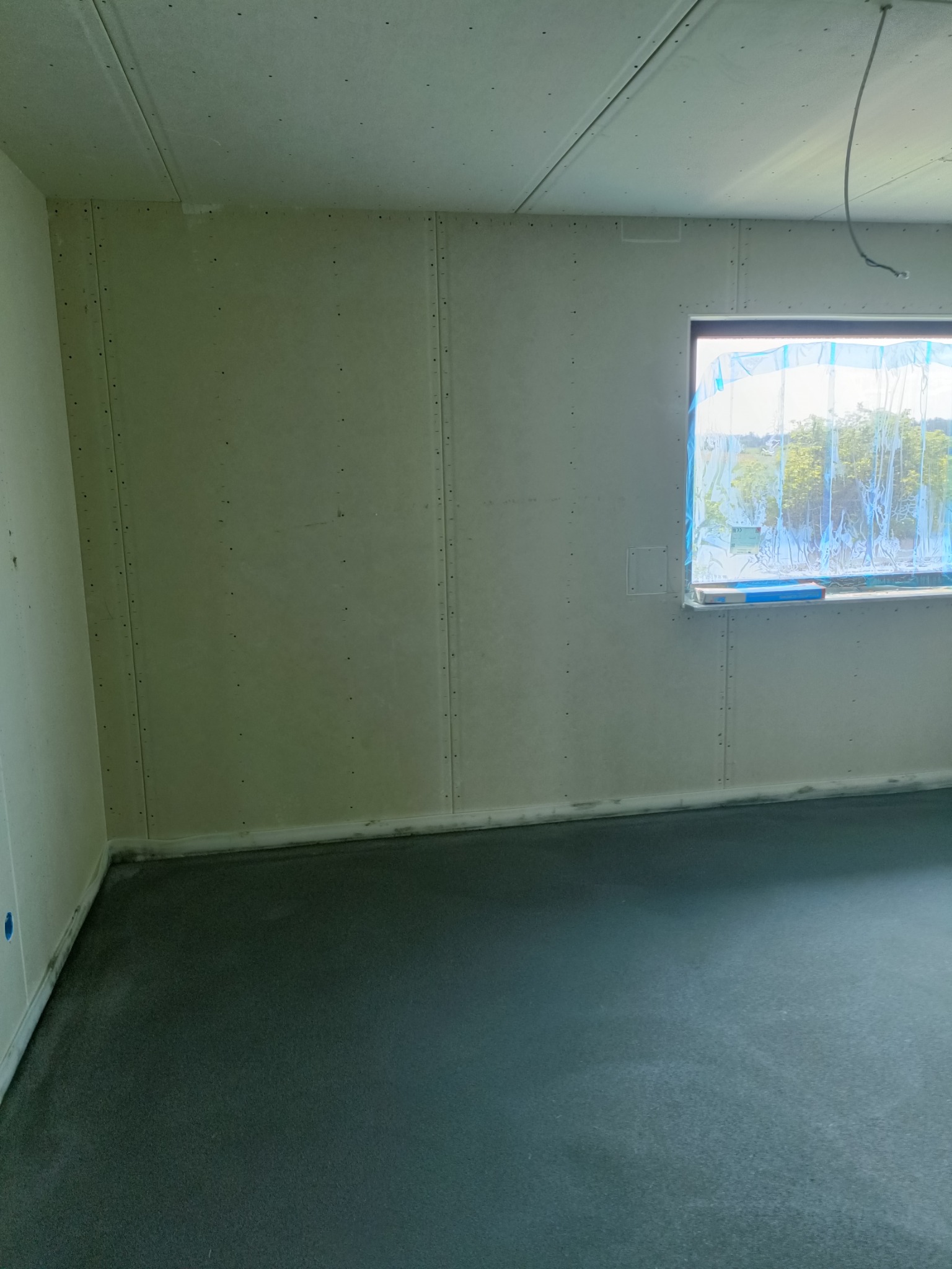 Surowe wnętrze w trakcie wykańczania: ściany i sufit pokryte płytami, zabezpieczone okno z widokiem na zieleń, wylewka na podłodze.