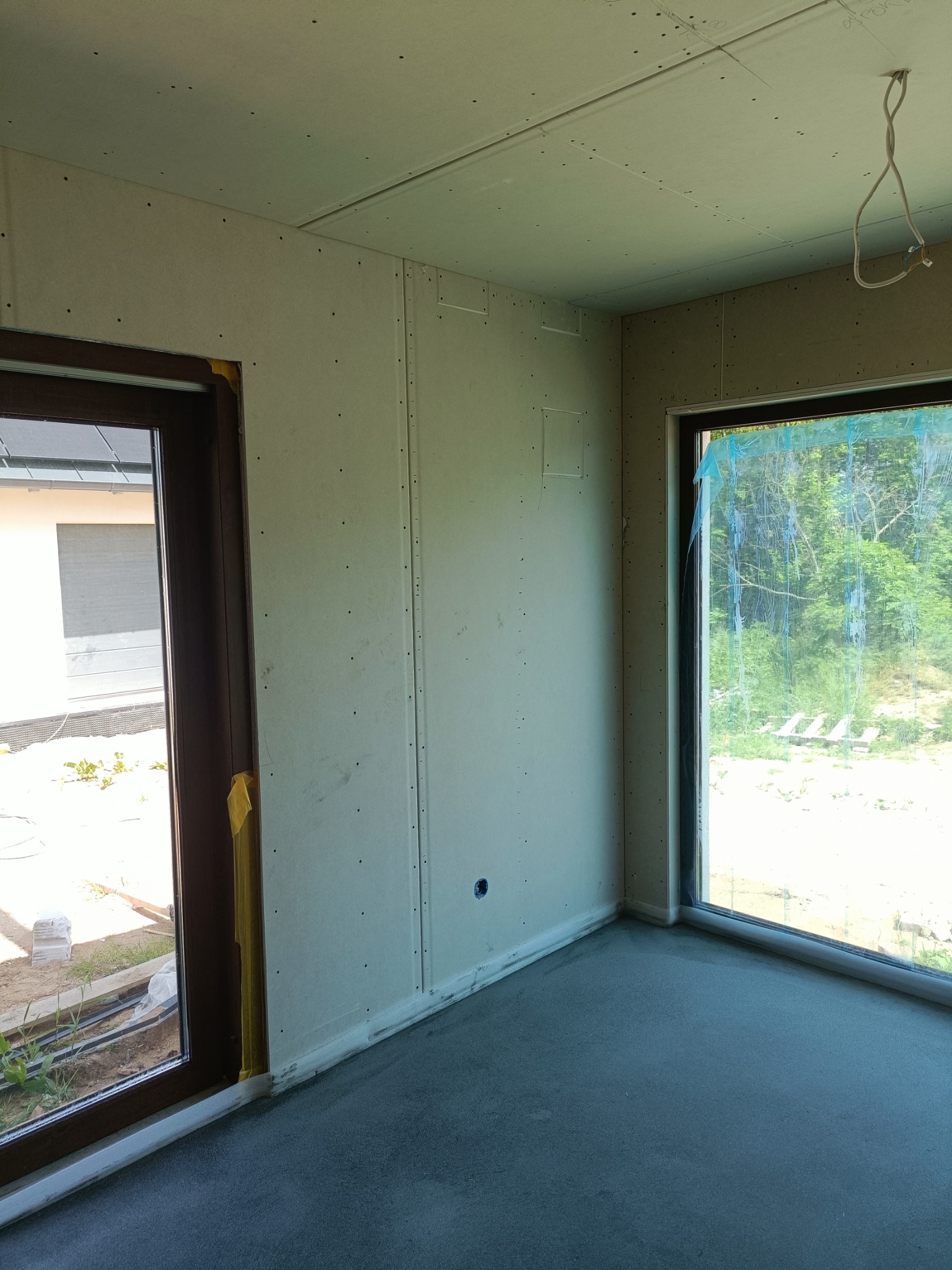 Wnętrze w trakcie prac wykończeniowych: ściany i sufit pokryte płytami gipsowo-kartonowymi, widoczne miejsca łączeń i otwory po wkrętach, surowa wylewka podłogowa, okna zabezpieczone folią...