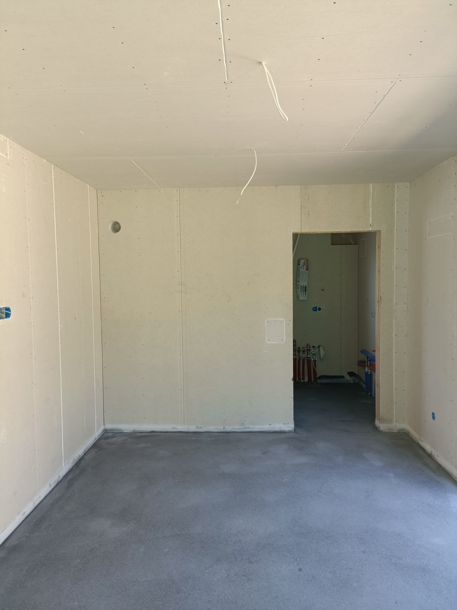 Surowe wnętrze w trakcie remontu: ściany wyłożone płytami gipsowo-kartonowymi, surowa wylewka na podłodze, widoczne instalacje w sąsiednim pomieszczeniu przez otwór drzwiowy.