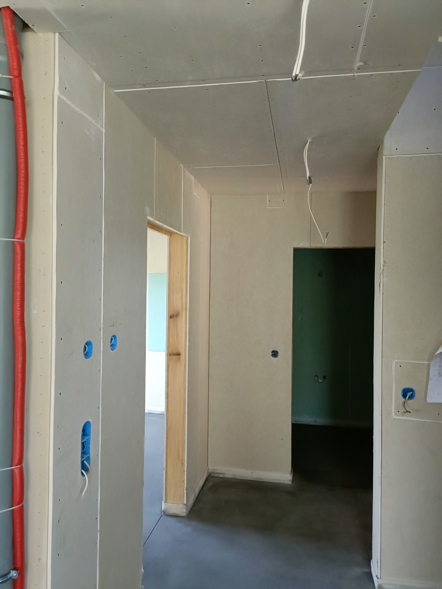Korytarz w trakcie prac wykończeniowych: ściany obłożone płytami gipsowo-kartonowymi, widoczne otwory pod instalacje elektryczne, czerwona rura instalacyjna przy ścianie, surowy beton na podłodze...