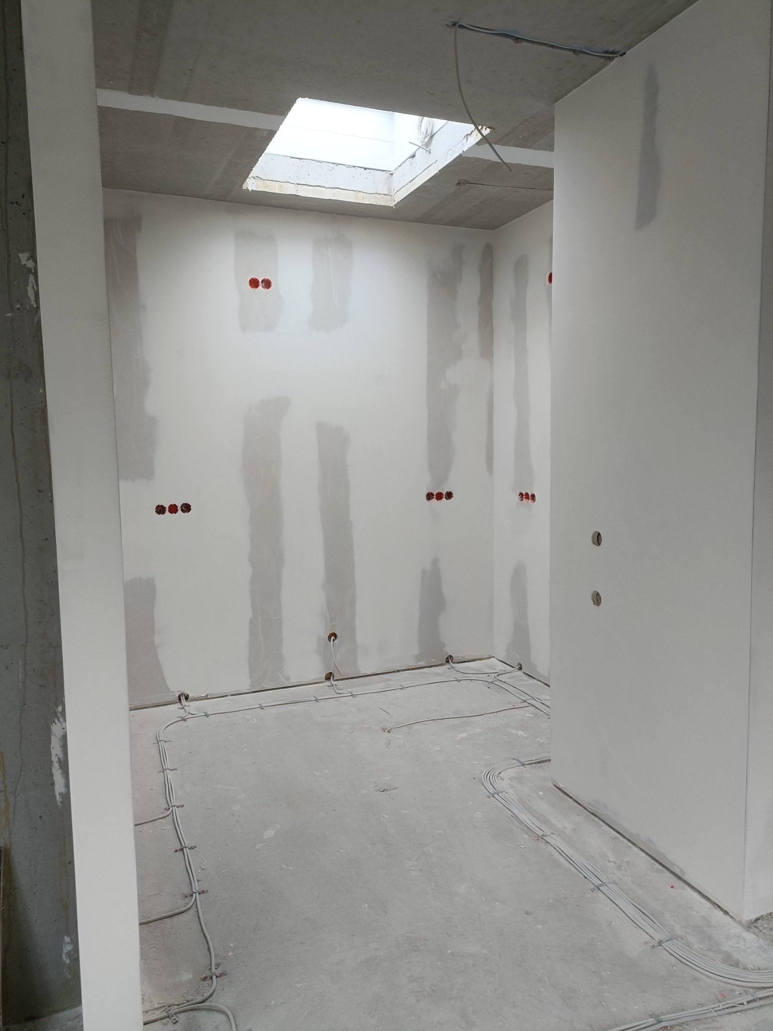 Wnętrze w trakcie prac wykończeniowych: ściany z widocznymi łączeniami płyt, zamontowane puszki elektryczne oraz kable ułożone na podłodze, widoczne okno dachowe.