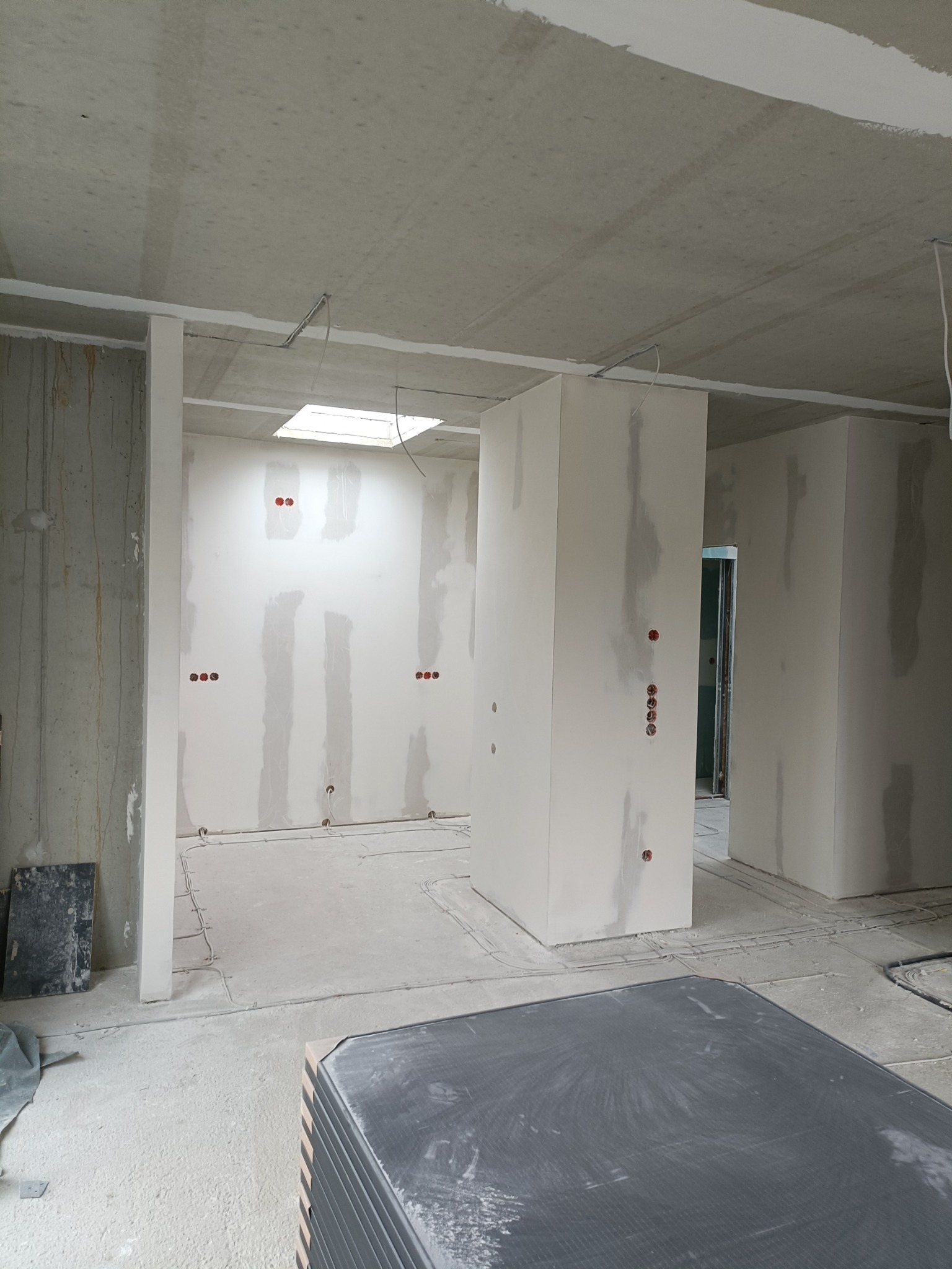 Wnętrze w trakcie prac wykończeniowych: surowy sufit, ściany z widocznymi łączeniami, wystające przewody elektryczne, otwór okienny w suficie i stos płyt na podłodze.