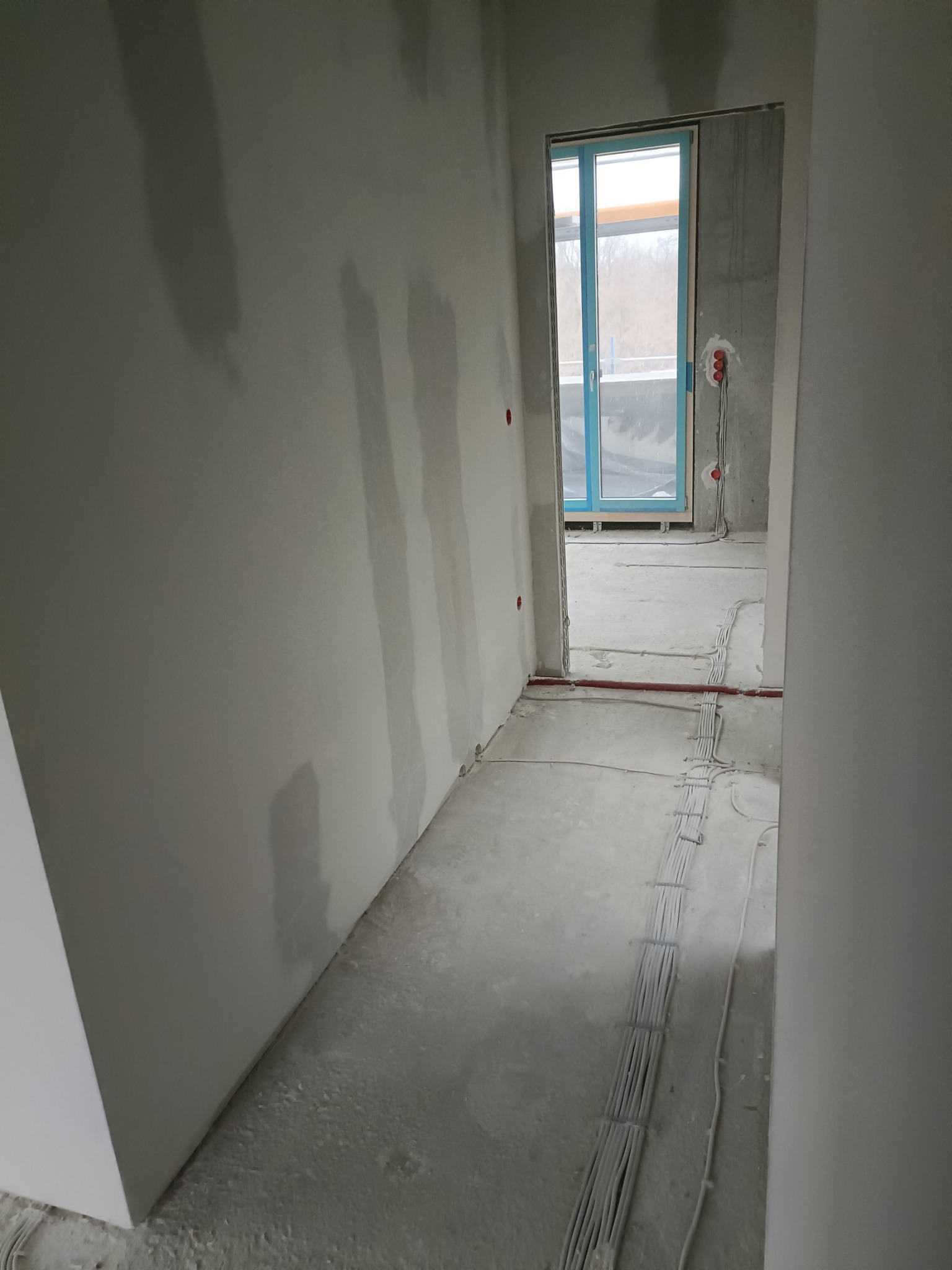 Korytarz w surowym stanie, widoczne przewody elektryczne ułożone wzdłuż ściany, ściany przygotowane pod malowanie, widoczne ślady po szpachlowaniu.