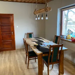 Drewniany stół z żywicą epoksydową w jadalni z drewnianą podłogą i lampą z pnia drzewa, widoczne narzędzia na blacie.