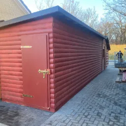 Czerwony domek narzędziowy z drewna, zamykany na złoty zamek, stoi na szarej kostce brukowej ułożonej we wzór jodełki, w tle widoczny człowiek z taczką i paleta z kostką.