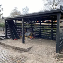 Drewniana wiata na narzędzia pomalowana na czarno, z widocznymi narzędziami ogrodniczymi i taczką w środku, ustawiona na kostce brukowej.