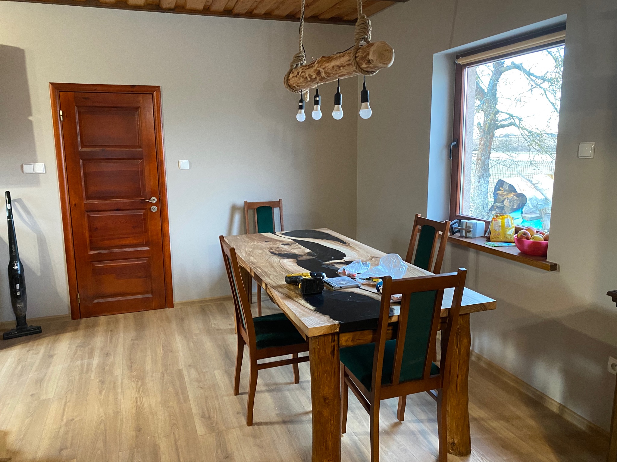 Drewniany stół z żywicą epoksydową w jadalni z drewnianą podłogą i lampą z pnia drzewa, widoczne narzędzia na blacie.