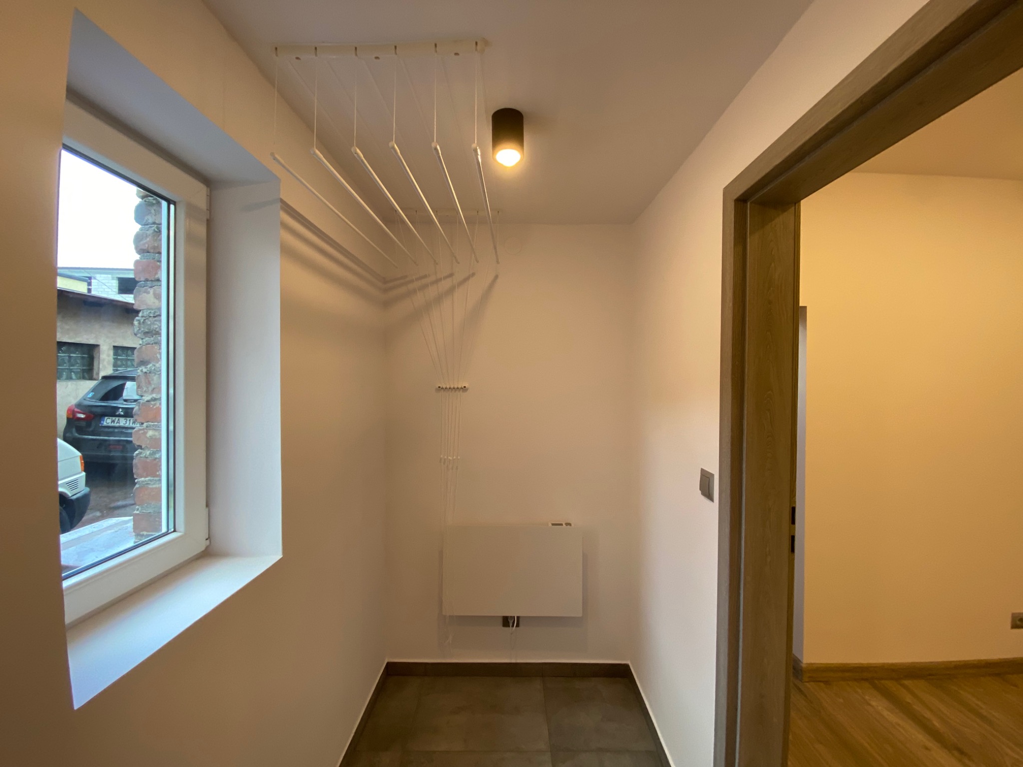 Wykończona piwnica w kamienicy: widok na pomieszczenie z szarą podłogą, suszarką sufitową, grzejnikiem na ścianie i oknem z widokiem na budynek z cegły i zaparkowany samochód.