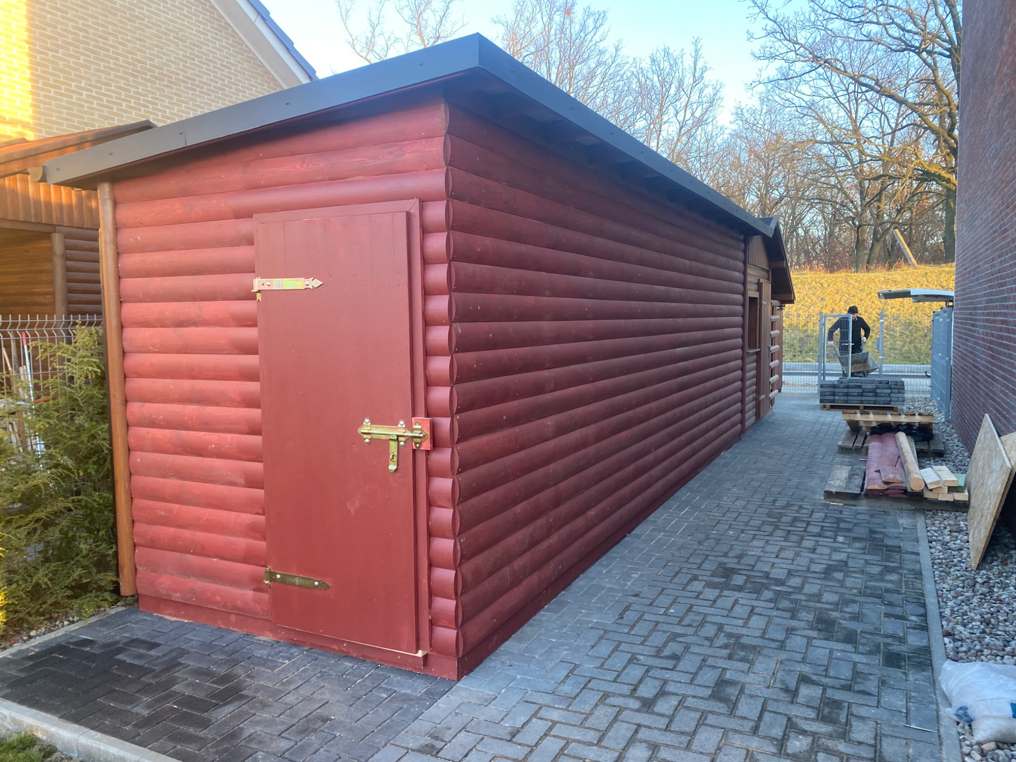 Czerwony domek narzędziowy z drewna, zamykany na złoty zamek, stoi na szarej kostce brukowej ułożonej we wzór jodełki, w tle widoczny człowiek z taczką i paleta z kostką.