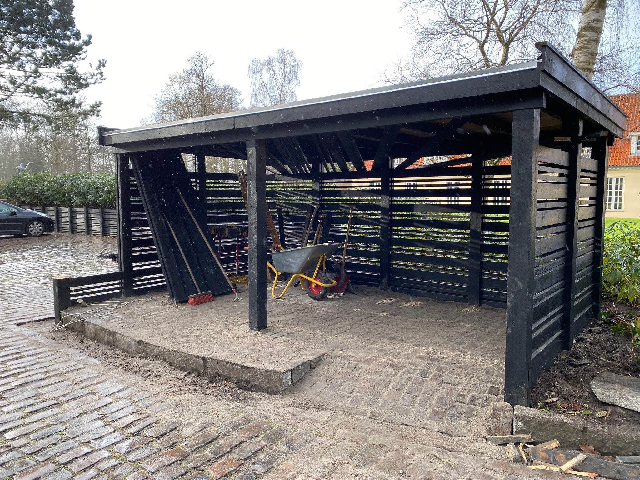 Drewniana wiata na narzędzia pomalowana na czarno, z widocznymi narzędziami ogrodniczymi i taczką w środku, ustawiona na kostce brukowej.