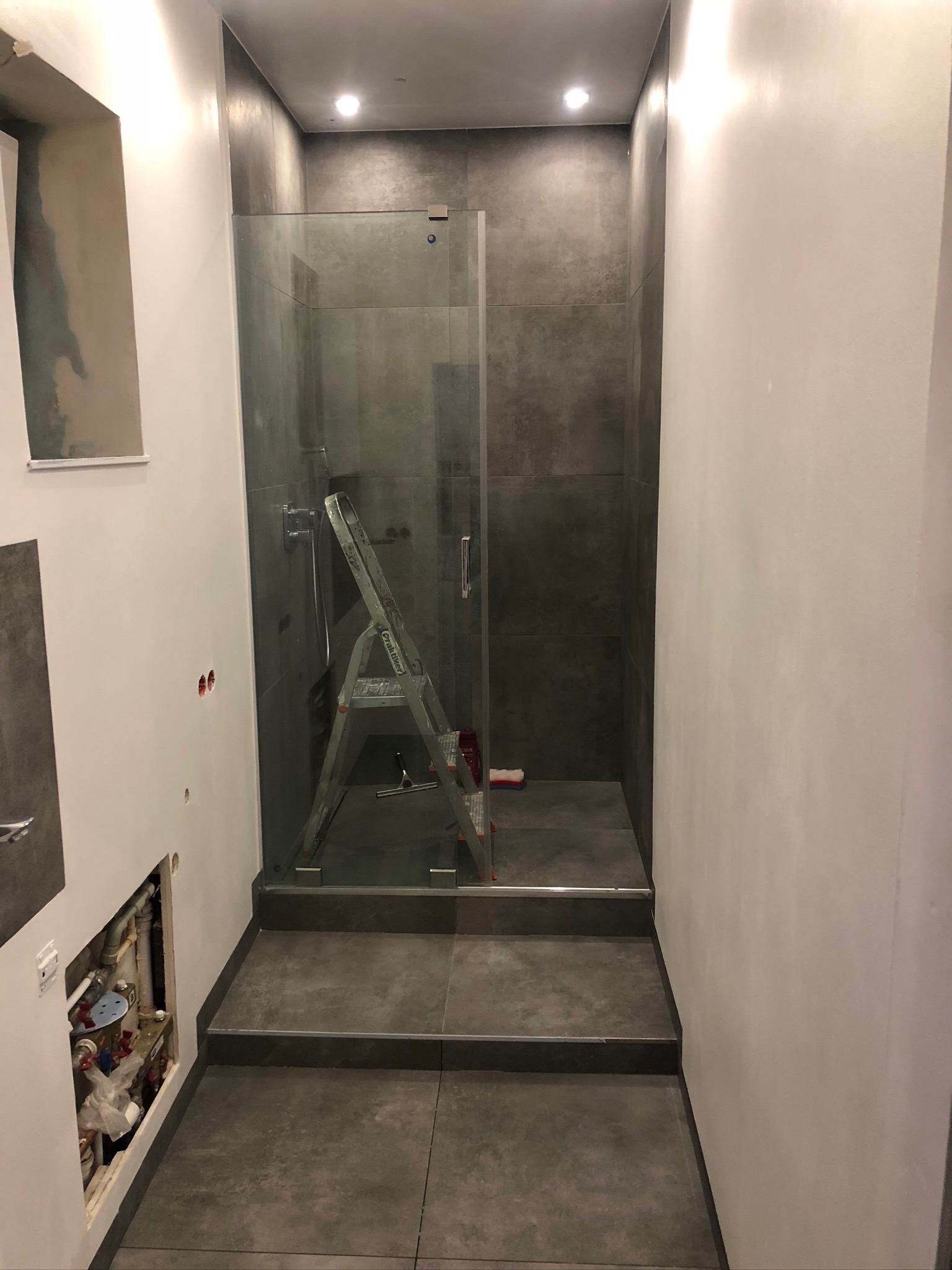 Wnętrze łazienki w trakcie prac wykończeniowych: widoczna kabina prysznicowa z ciemnymi płytkami, drabina oparta o ścianę kabiny, odkryte rury instalacji wodnej w ścianie oraz ściany przygotowane...