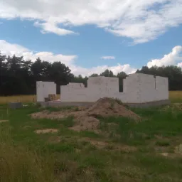 Wznoszone ściany parteru budynku z betonu komórkowego na fundamencie, widoczna paleta z bloczkami, teren budowy z wysoką trawą i stertą ziemi, w tle las i błękitne niebo z chmurami.