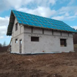 Wznoszony dwukondygnacyjny budynek z białych bloczków, z niebieską membraną dachową i drewnianą konstrukcją dachu, na placu budowy z widoczną drabiną opartą o ścianę boczną.