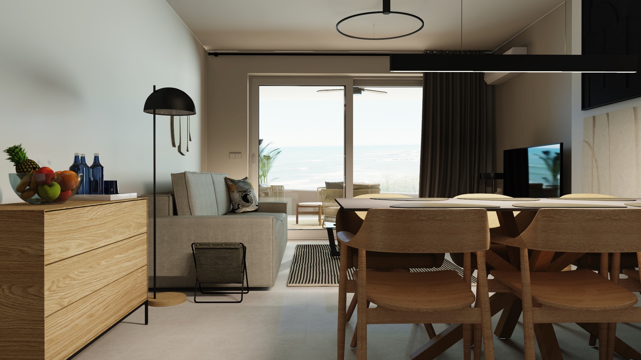 Jasne wnętrze apartamentu z widokiem na morze: drewniana komoda z owocami, szara sofa z poduszką, stół jadalny z krzesłami z jasnego drewna, lampa podłogowa i czarne akcenty oświetleniowe.