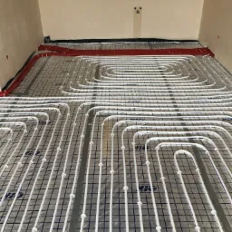 Białe rury ogrzewania podłogowego ułożone spiralnie na folii z siatką w surowym pomieszczeniu z widocznymi śladami po instalacjach na ścianach.