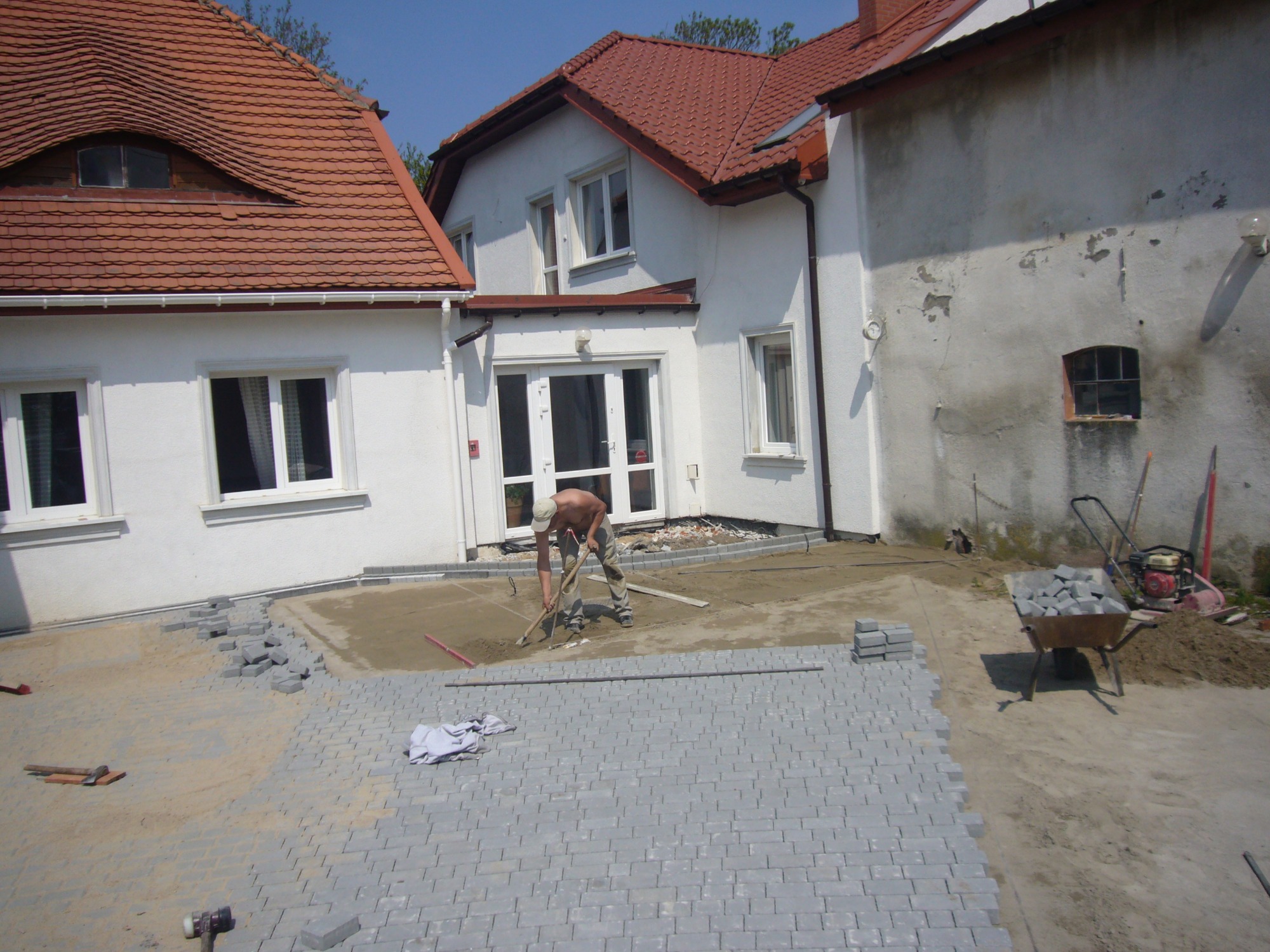 Mężczyzna w czapce wyrównuje podłoże pod szarą kostkę brukową na posesji przed domem z czerwoną dachówką; w tle taczka z kostką i zagęszczarka.