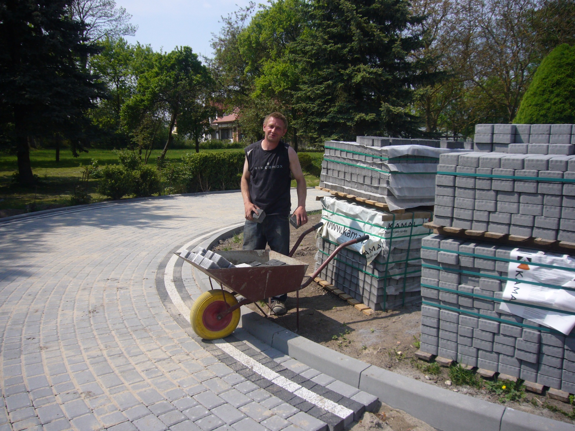 Pracownik z taczką pełną kostki stoi na nowo ułożonym, zakrzywionym chodniku z kostki, obok palet z kostką i krawężników.