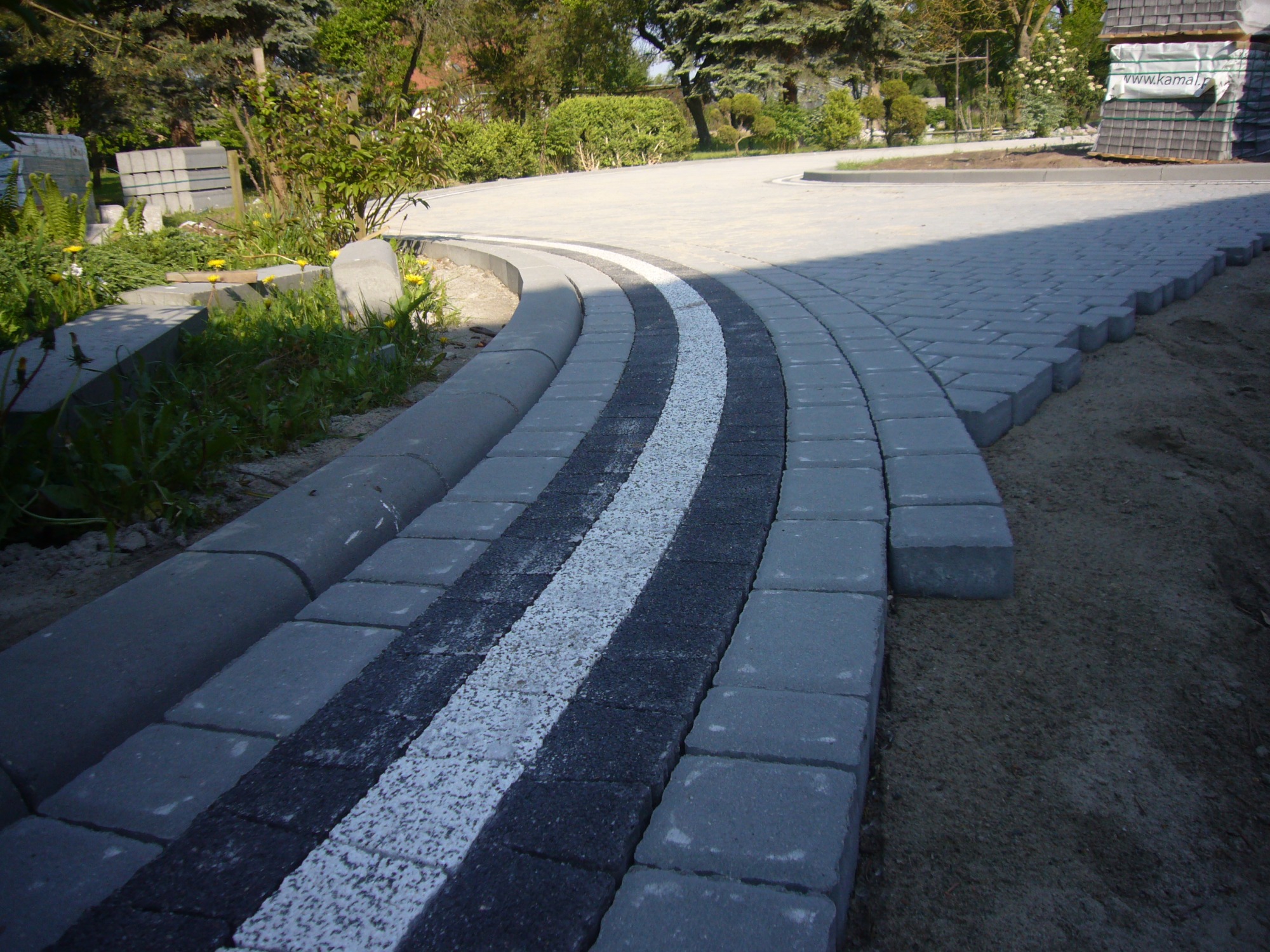 Zakrzywiony podjazd z szarej kostki brukowej z białym pasem dekoracyjnym, obrzeża z kostki betonowej, widoczne niezakończone prace i przygotowane elementy.
