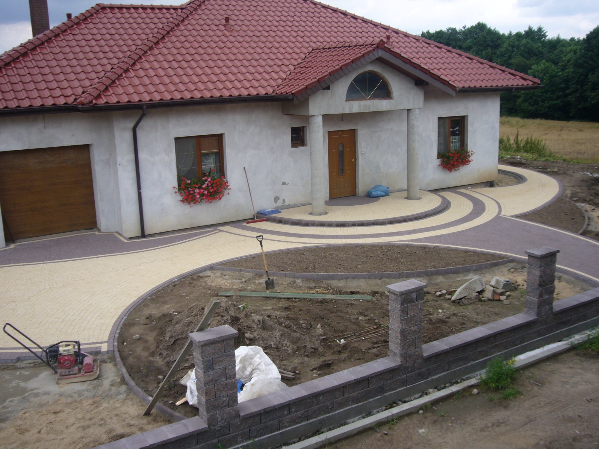 Układanie kostki brukowej na podjeździe domu z elementami dekoracyjnymi, widoczny fragment budowanego murku z bloczków kamiennych i zagęszczarka w tle.