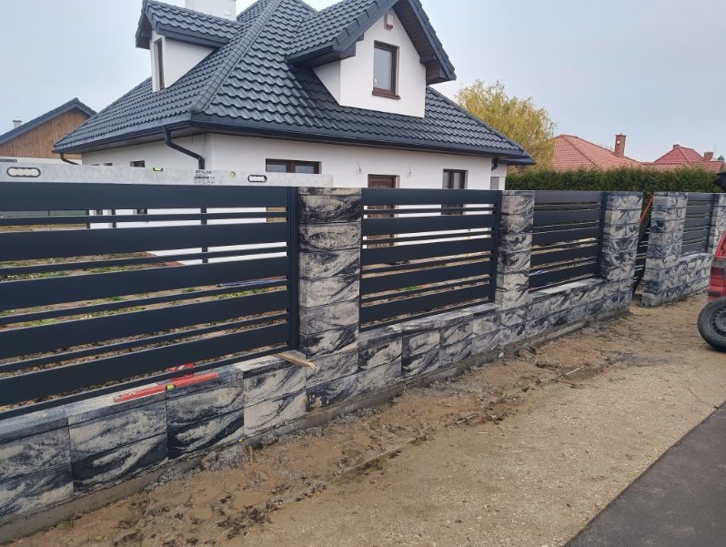 Nowoczesne ogrodzenie panelowe z elementami imitującymi kamień, poziome przęsła w kolorze antracytowym, widoczne poziomice, dom w tle.