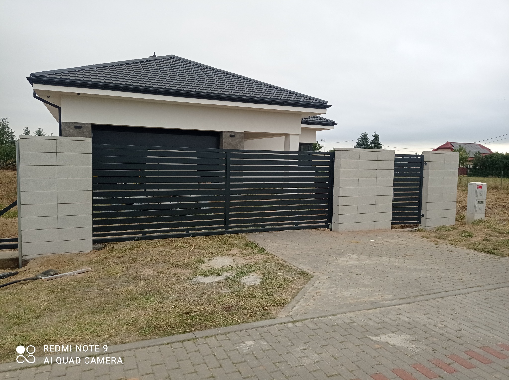 Nowoczesna, pozioma brama wjazdowa z czarnymi przęsłami i szarymi słupkami z bloczków betonowych, na tle domu z ciemną dachówką i szarą kostką brukową.