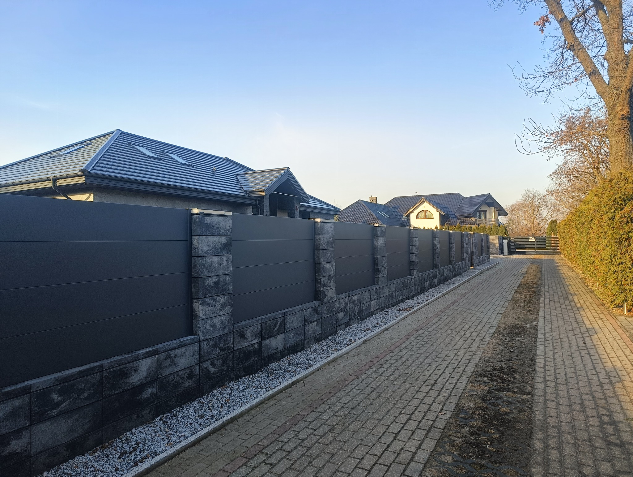 Nowoczesne ogrodzenie panelowe z podmurówką z szarej kostki, widok z perspektywy chodnika, domy w tle, słoneczny dzień.