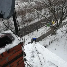 Pracownik w niebieskiej kurtce na zasypanym śniegiem dachu podczas opadów śniegu, widok z góry, obok komin z cegły i antena satelitarna.