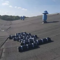Czarny dach pokryty papą termozgrzewalną w rolkach, przygotowany do montażu, z niebieskimi kominkami wentylacyjnymi w tle pod błękitnym niebem.