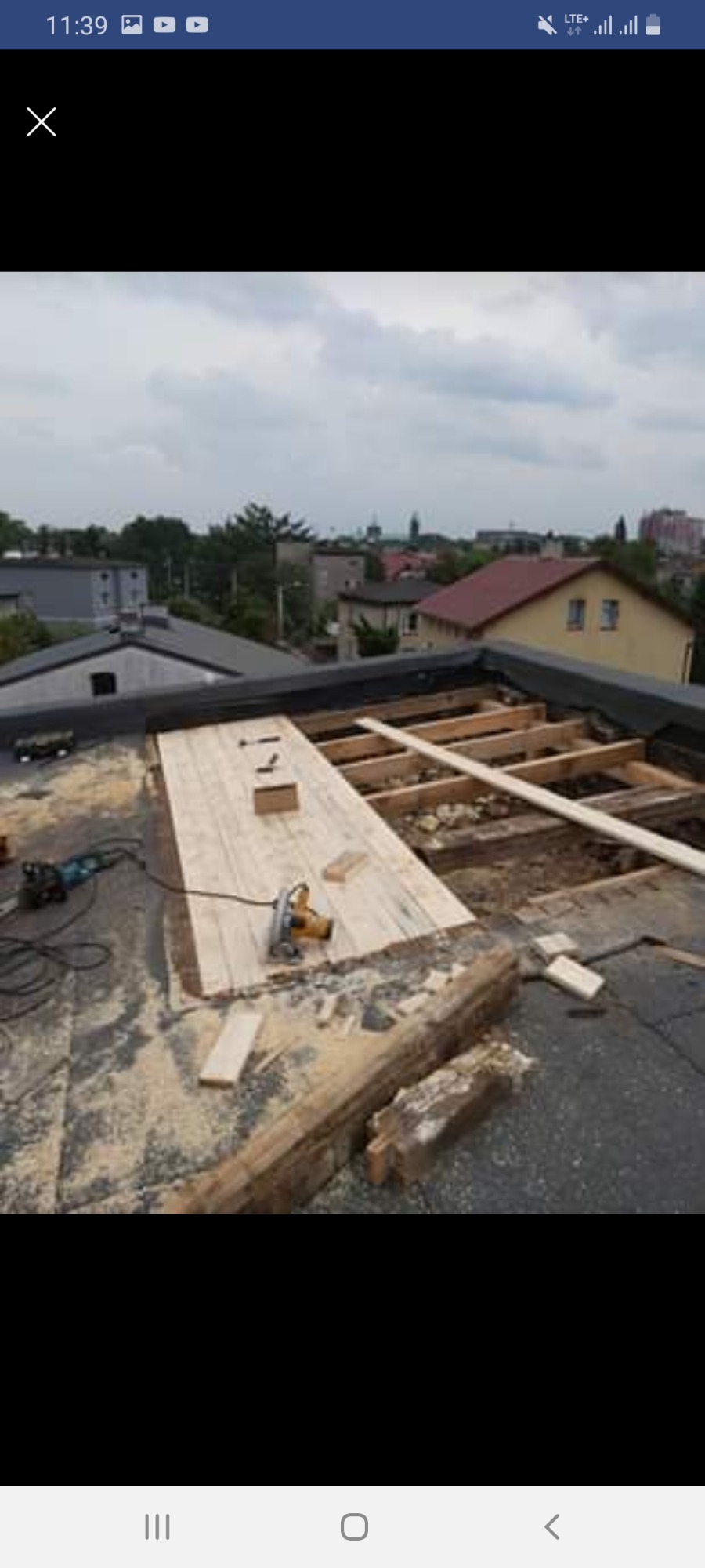 Prace dekarskie w toku: widok z góry na płaski dach z odsłoniętą konstrukcją drewnianą, częściowo pokryty płytami, z widoczną piłą elektryczną i narzędziami, w tle zabudowa miejska pod pochmurnym...