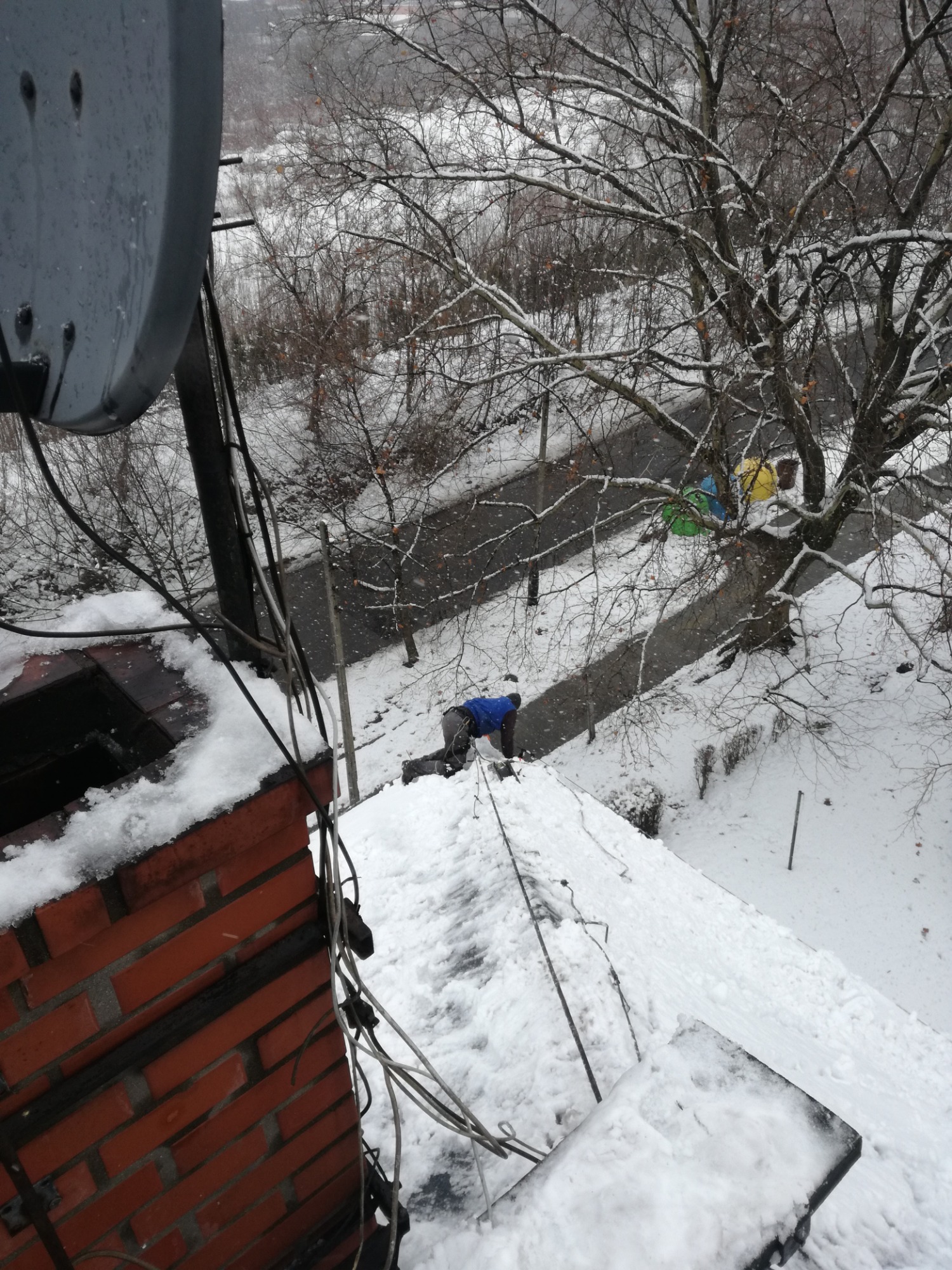 Pracownik w niebieskiej kurtce na zasypanym śniegiem dachu podczas opadów śniegu, widok z góry, obok komin z cegły i antena satelitarna.