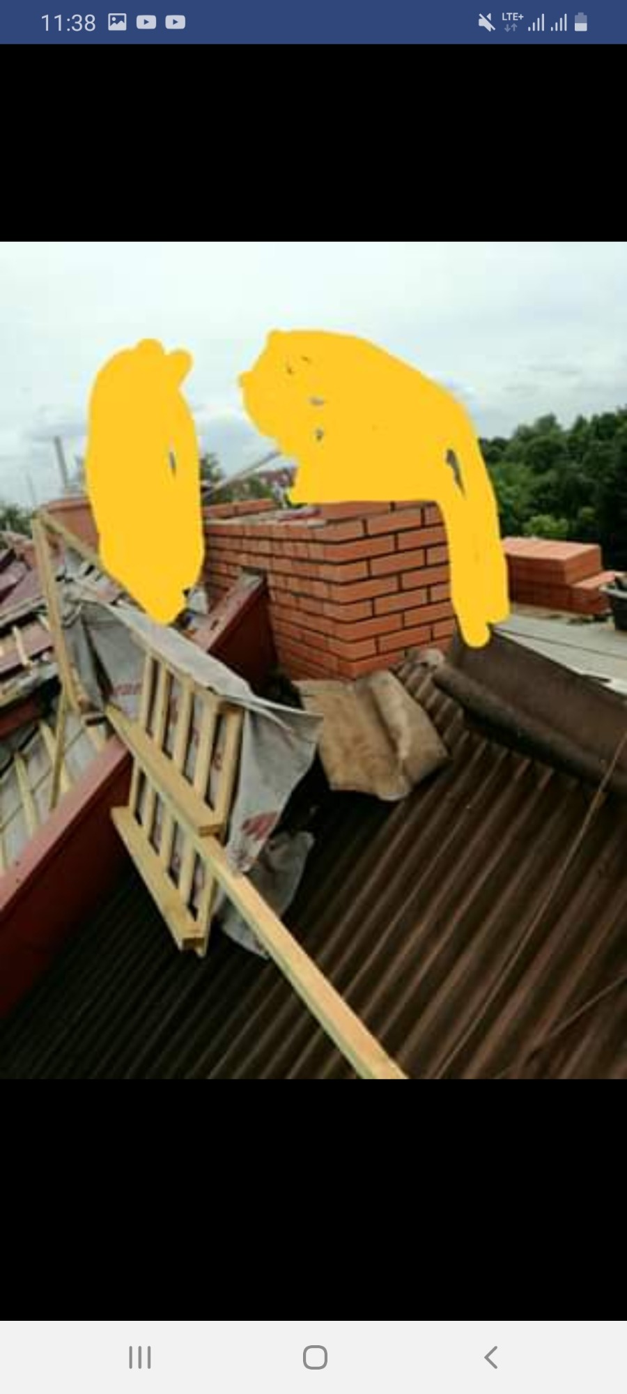 Rozłożona drabina na dachu z brązowej blachy trapezowej w trakcie budowy komina z czerwonej cegły, widok na zielone drzewa w tle pod pochmurnym niebem.