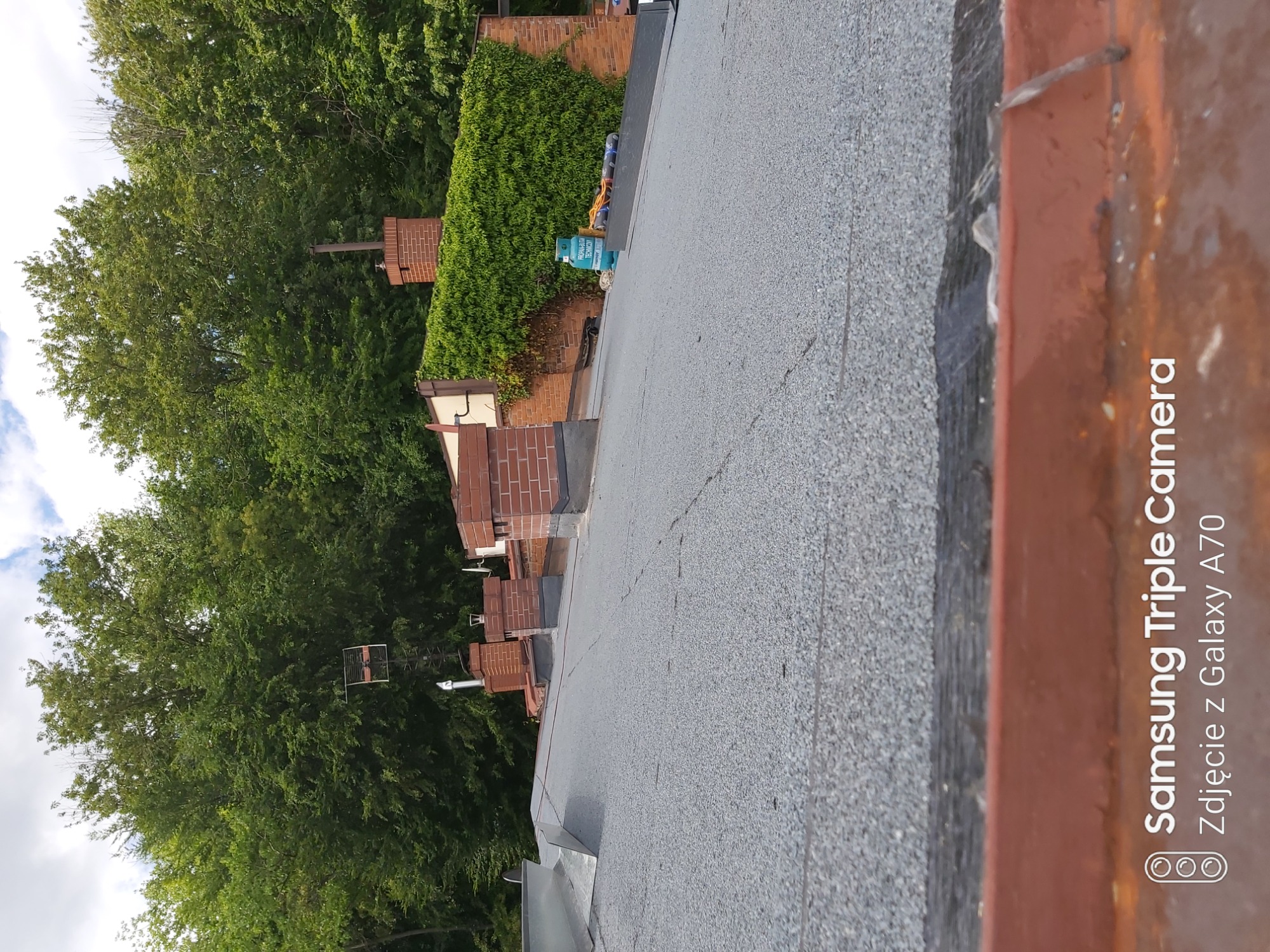 Nowe pokrycie dachowe z papy bitumicznej na tle zieleni, widoczne kominy i butla gazowa z palnikiem dekarskim.