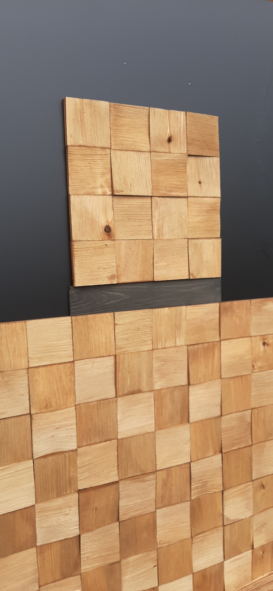 Fragment dachu z drewnianych kwadratowych gontów ułożonych szachownicę, z widocznym sękiem na jednym z elementów, na szarym tle ściany.