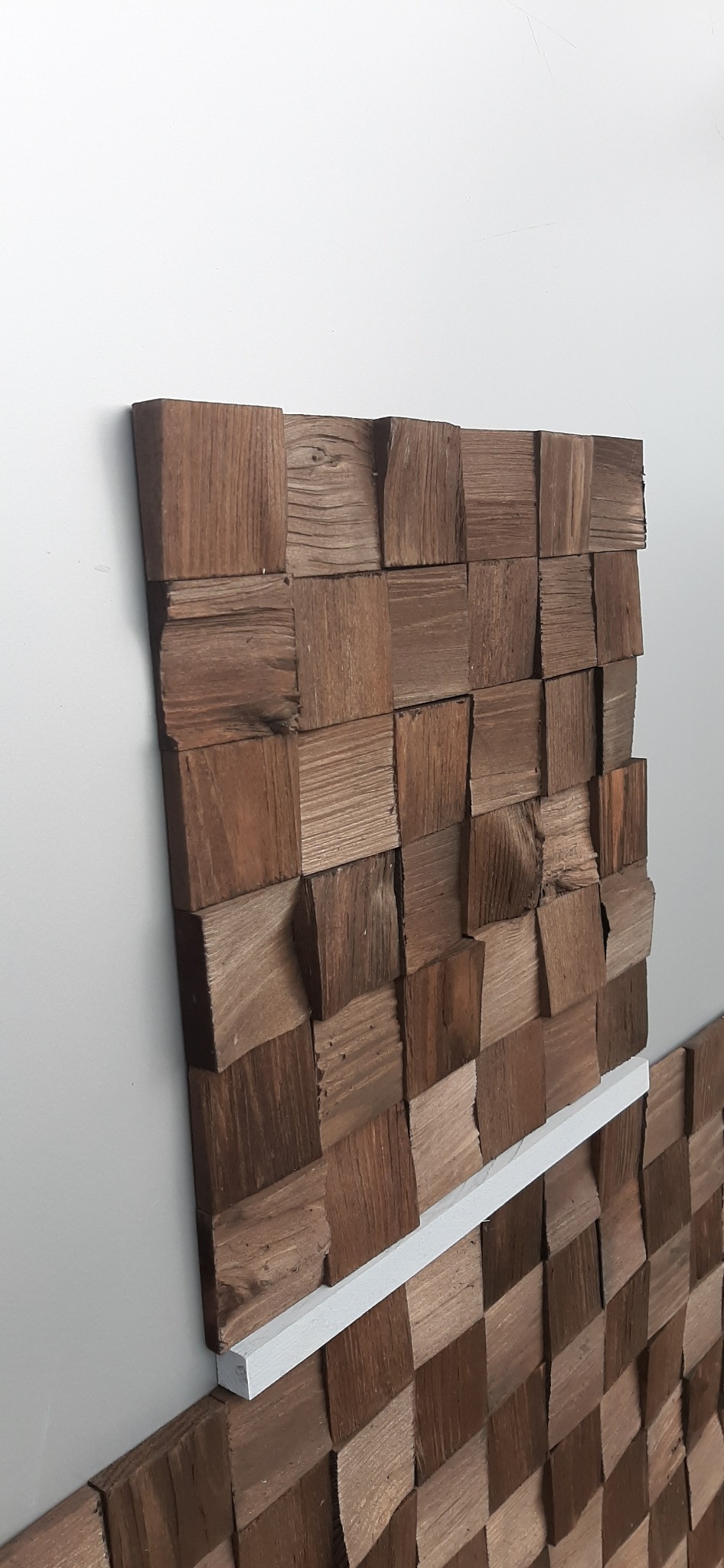 Dekoracyjna ściana z drewnianych kostek o zróżnicowanej fakturze i odcieniach brązu, zamontowana na białej ścianie, z widoczną białą listwą przypodłogową.