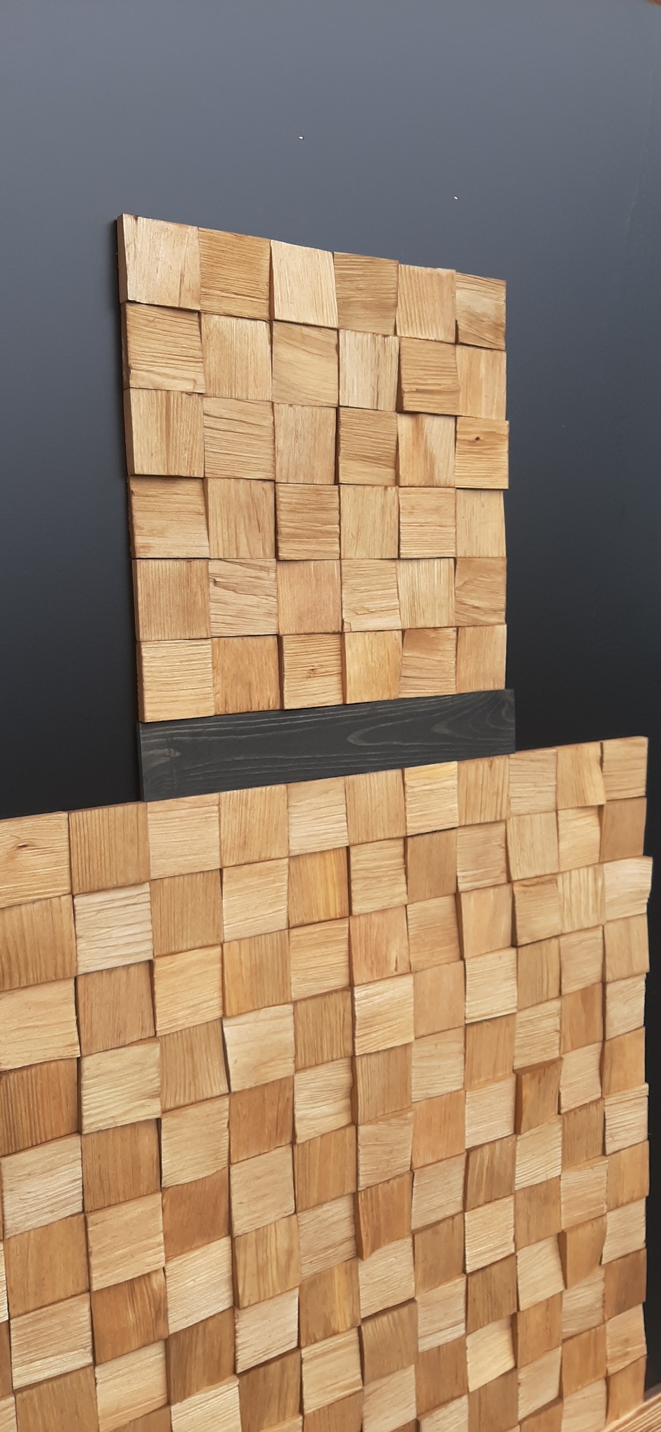 Dekoracyjna ściana z drewnianych kostek o ciepłym odcieniu, ułożonych w szachownicę z elementem czarnej listwy, na tle ciemnoszarej ściany.