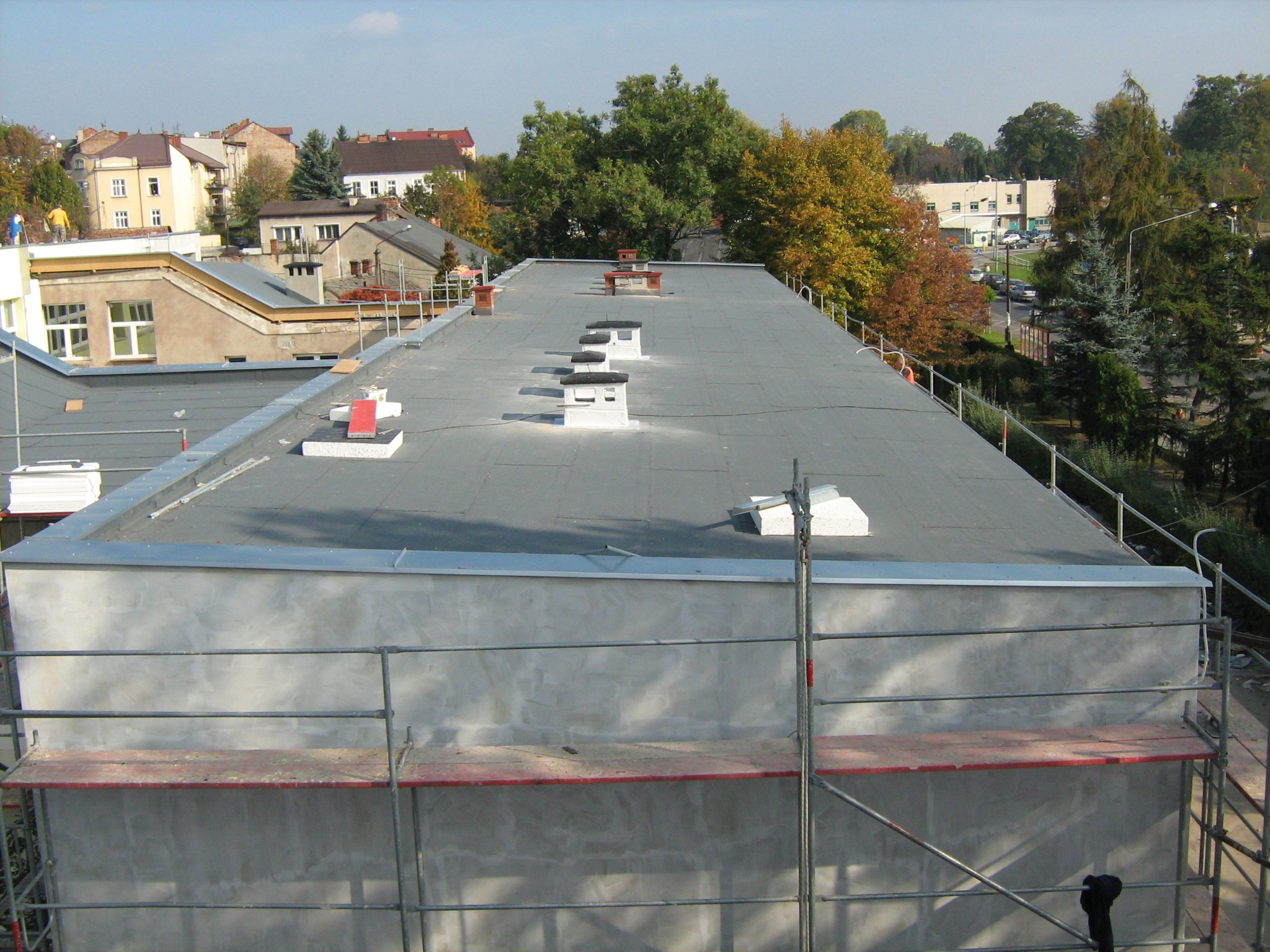 Płaski dach pokryty papą termozgrzewalną, widoczne kominki wentylacyjne i obróbki blacharskie, w tle zabudowa miejska i zieleń, rusztowanie przy attyce.